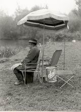 Auf dem Bild ist ein Angler bei relativ trübem Wetter zu sehen. Zum Schutz vor Regen hat er einen Sonnenschirm aufgestellt.